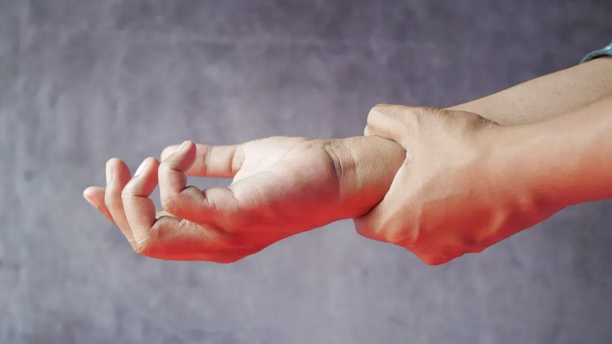 Dos manos en primer plano: una sujeta la muñeca de la otra, con la zona enrojecida, sobre un fondo gris; ilustra el dolor en las articulaciones
